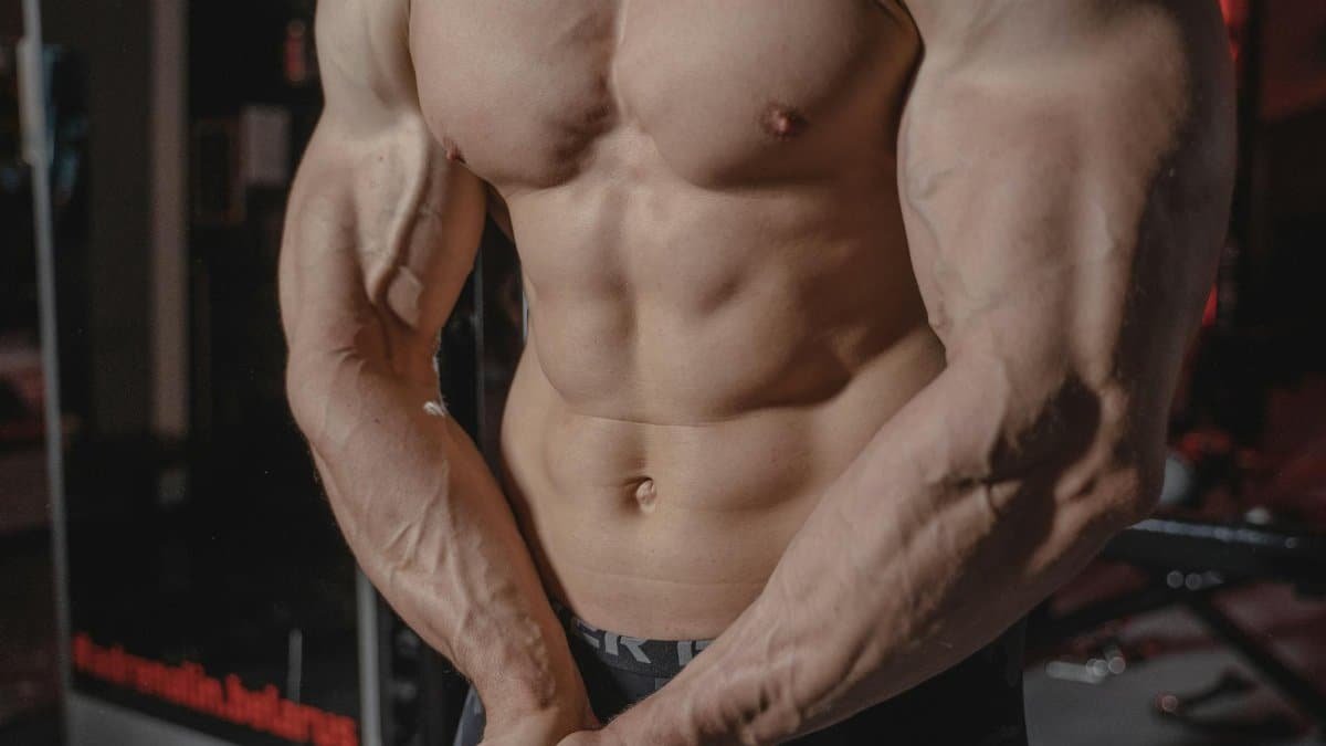 Shirtless male bodybuilder showing muscular torso and defined arms in a gym setting.