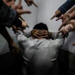 A person crouches in a corner surrounded by pointing fingers, illustrating social pressure and bullying.