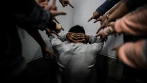 A person crouches in a corner surrounded by pointing fingers, illustrating social pressure and bullying.
