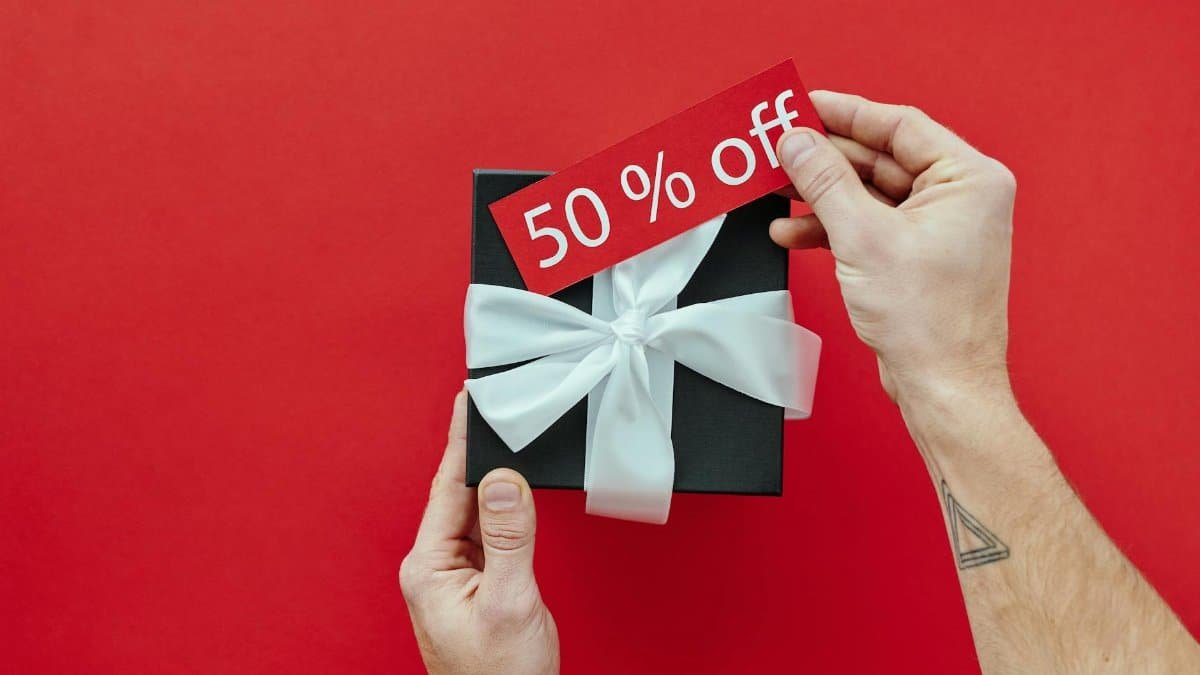 Black gift box with white ribbon held on red background, featuring a 50% off sale tag.