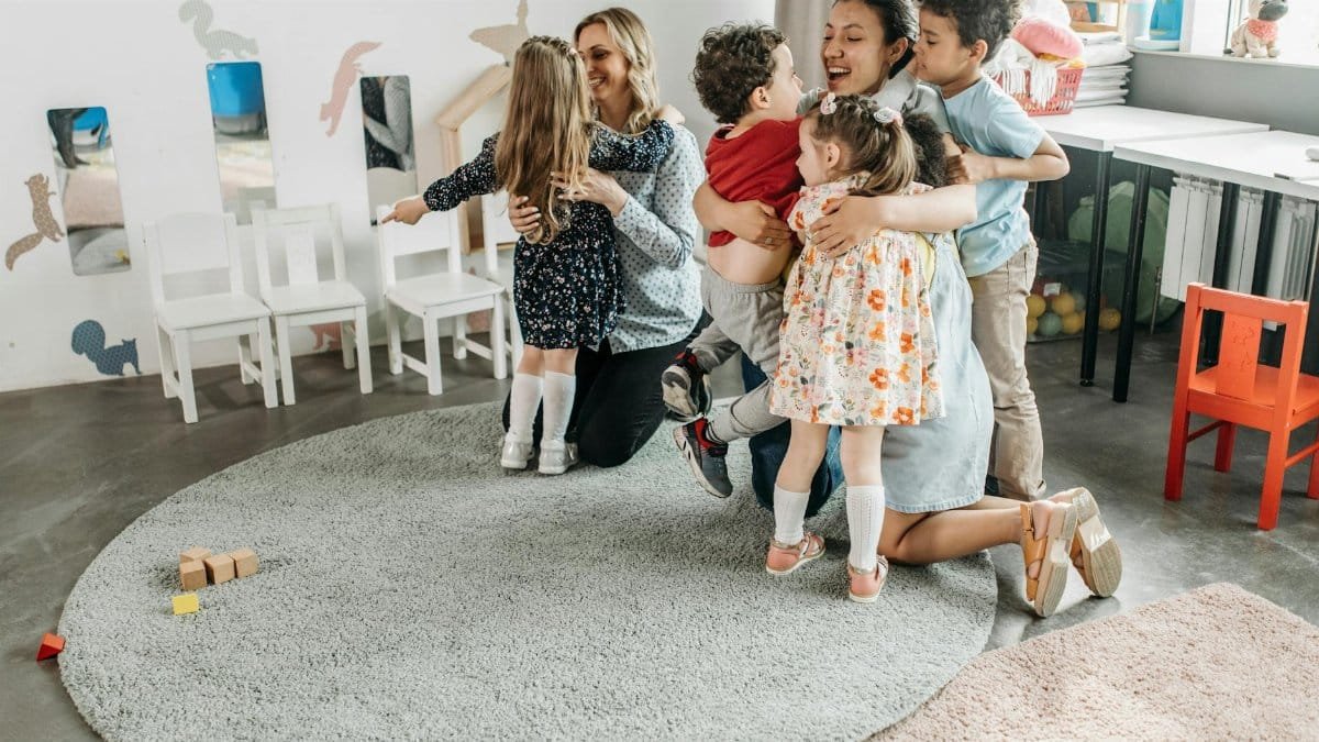 A group of children happily hugging their teachers in a kindergarten classroom.