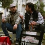 Two men converse at an outdoor flea market, surrounded by goods and greenery.