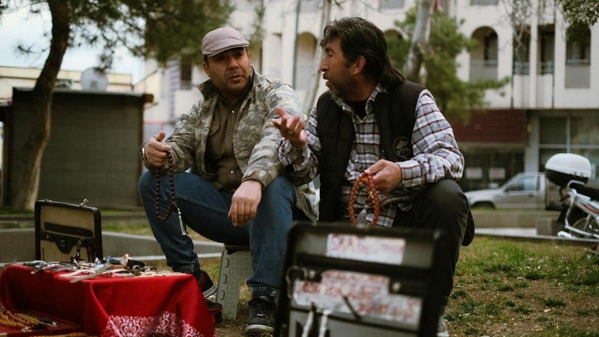 Two men converse at an outdoor flea market, surrounded by goods and greenery.