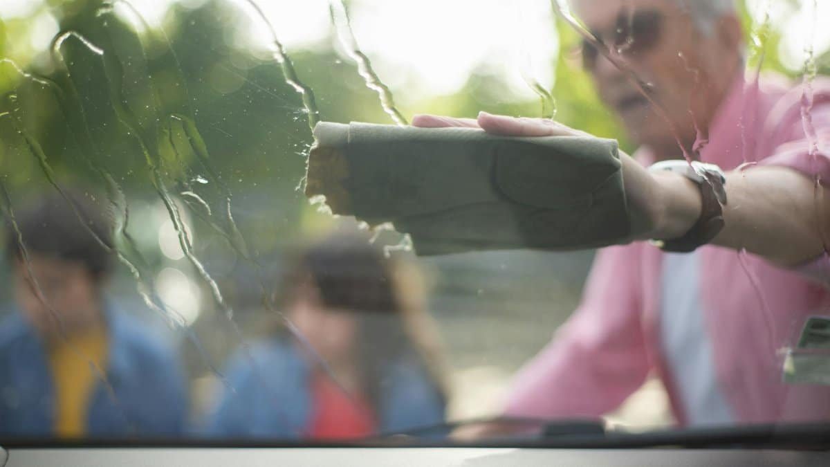 An elderly man cleans a car windshield with a cloth outdoors, capturing a moment of maintenance and care.