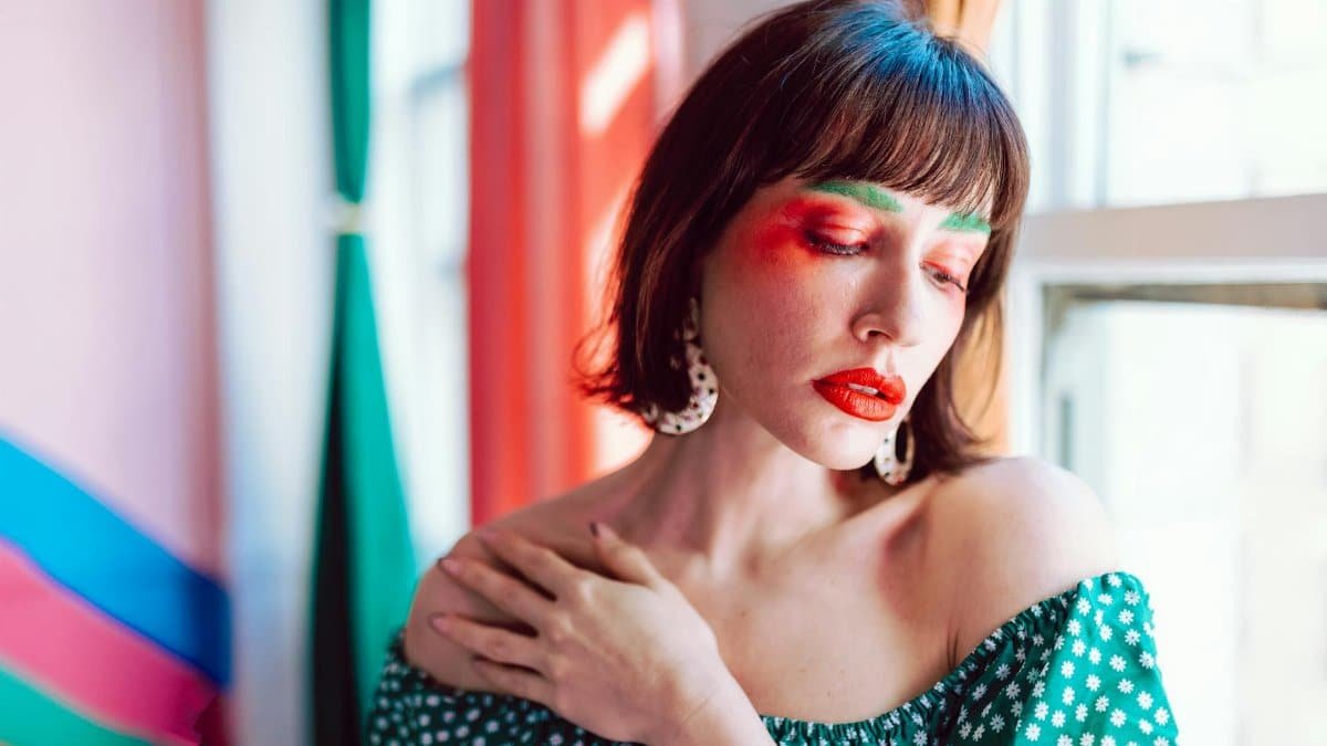 Colorful portrait of a woman in floral off-shoulder top with vibrant makeup near window.