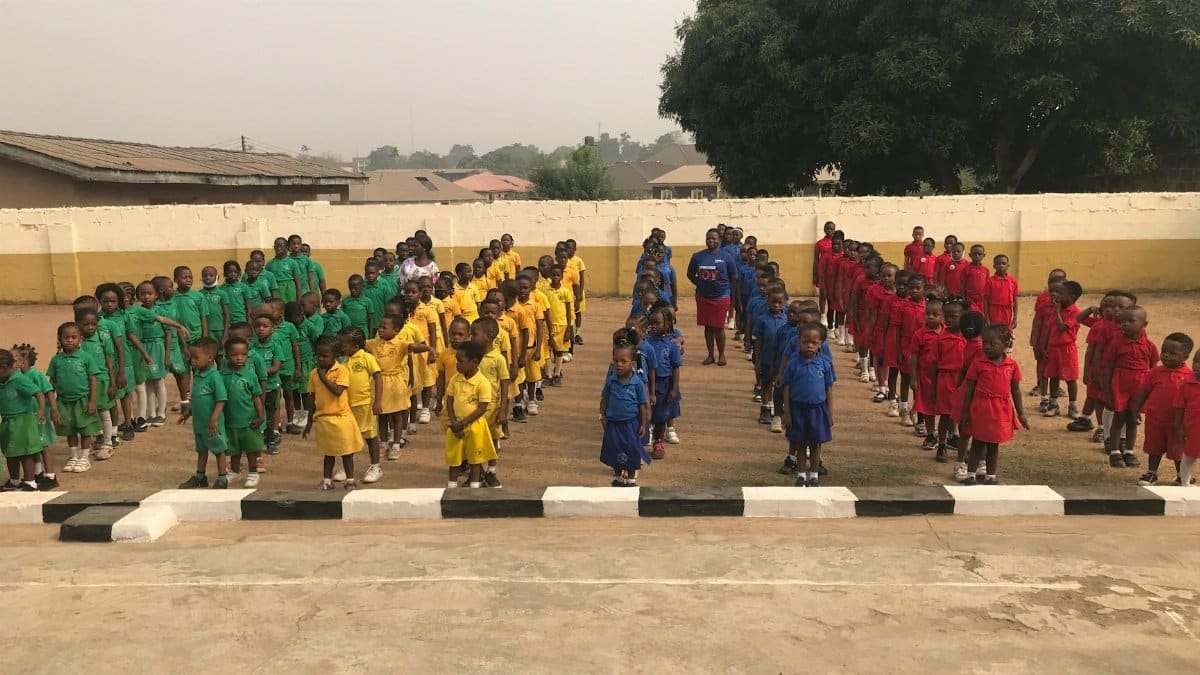 Children in colorful uniforms stand in formation on a school ground for assembly.