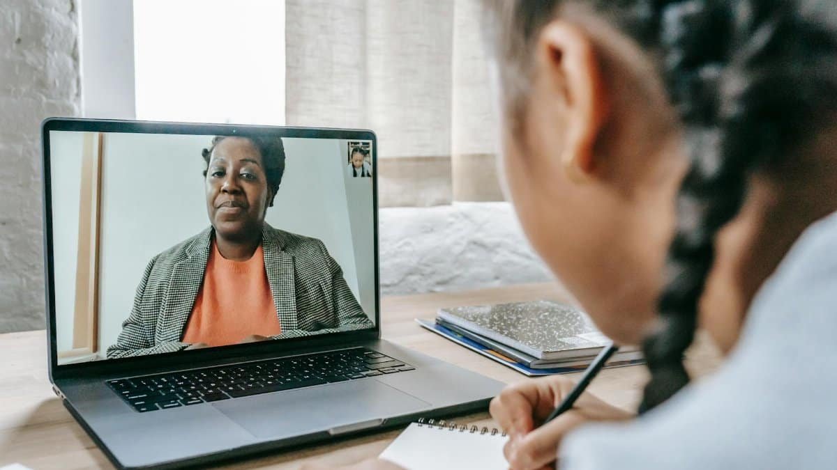A girl participating in an online learning session with a tutor via video call on her laptop, taking notes.