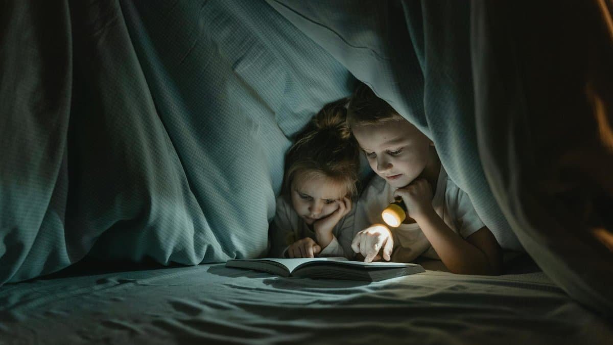Two children read a book under a blanket using a flashlight, showcasing a cozy night in.