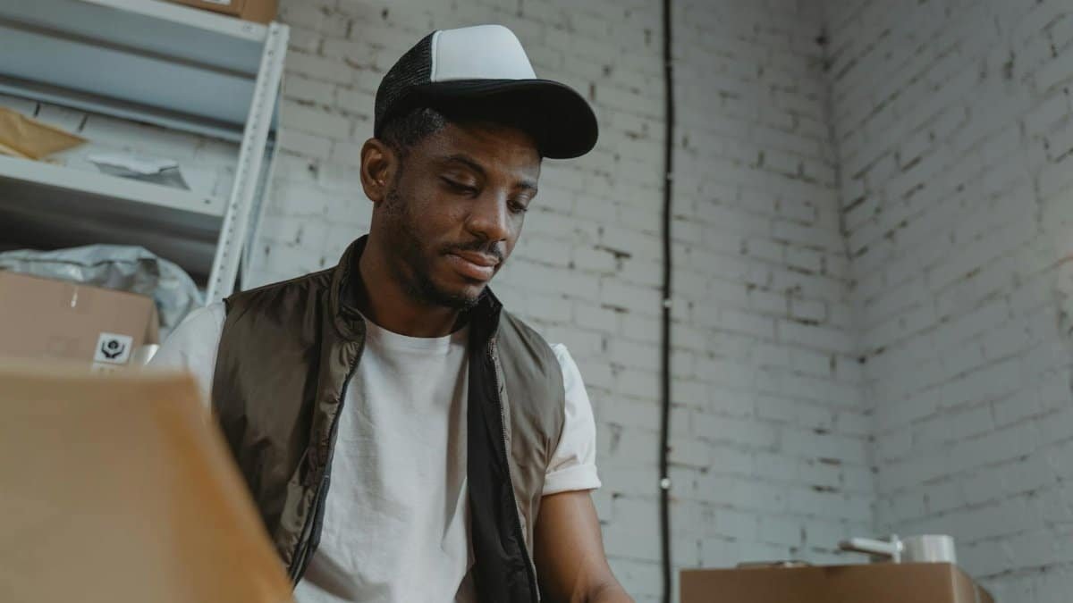 Man in a cap organizing packages in a warehouse with brick walls.