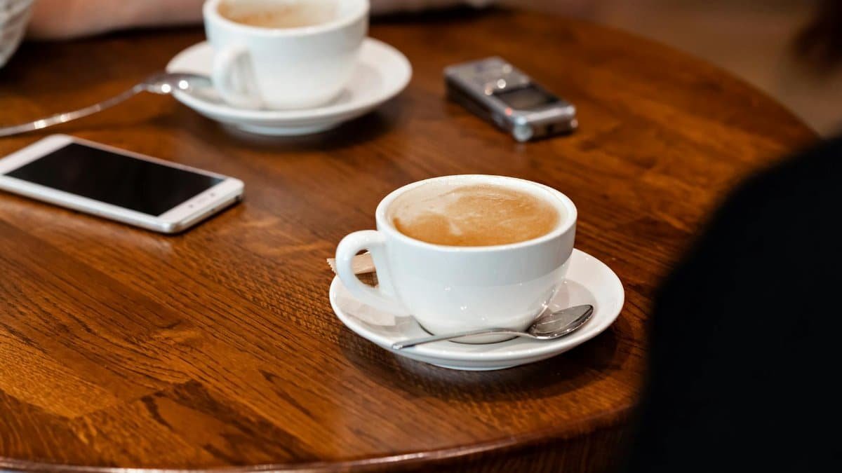 High angle of mugs with latte on saucer with spoon placed on wooden table near phone and voice recorder near crop anonymous person in light cafeteria