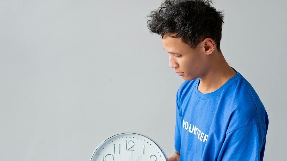 A young man in a blue shirt with 'Volunteer' on it holds a wall clock. Time concept.
