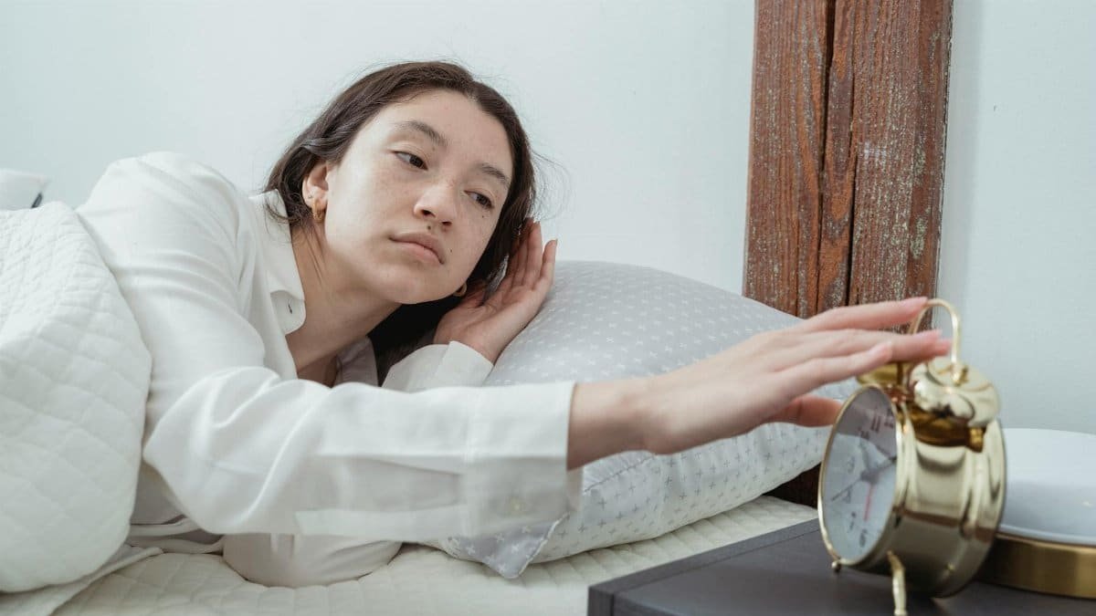 Sleepy young brunette female in white sleepwear resting in comfortable bed and waking up with alarm clock at home in morning time