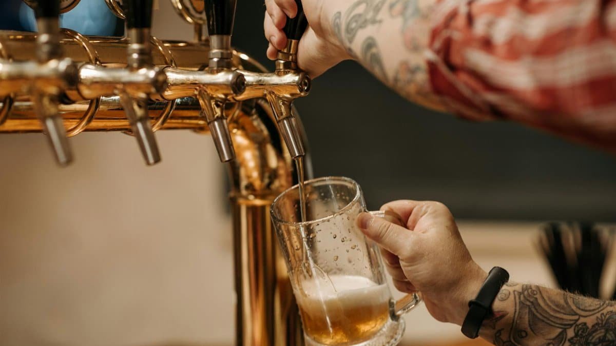 Tattooed bartender pouring a pint of beer from a golden tap, close-up view at a bar.