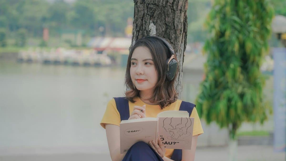 Woman enjoying a book under a tree while listening to music in a peaceful park setting.