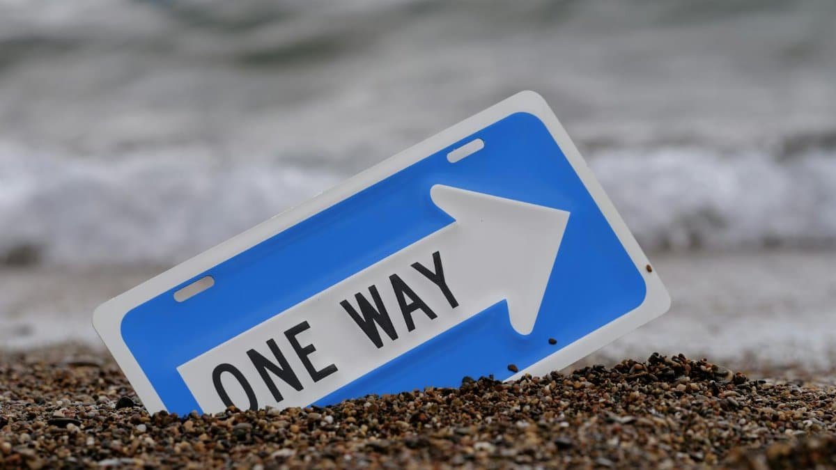A blue one way sign in the sand by the ocean, with waves in the background.
