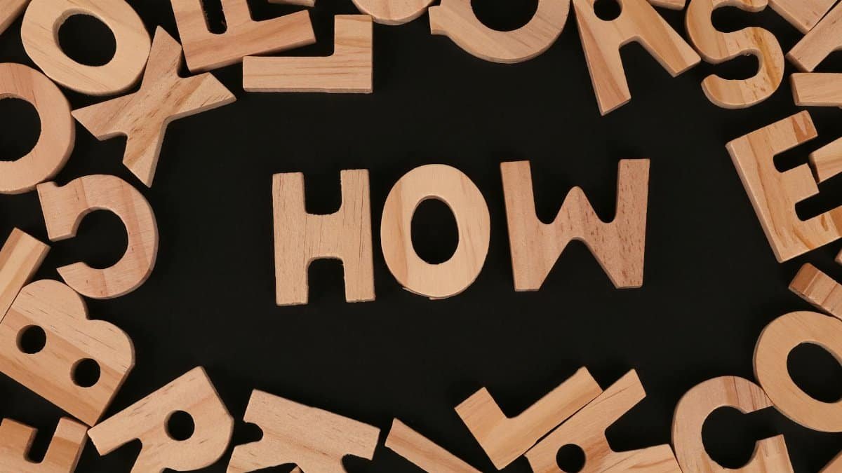 Top view of scattered wooden letters with 'HOW' centered on a dark surface, promoting inquiry.