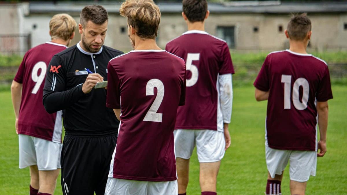 Soccer referee taking notes as team gathers on the field for a match outdoors.