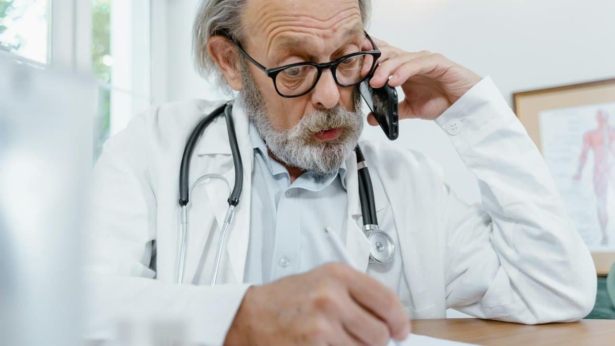 Elderly doctor in office on phone call with patient, writing notes.