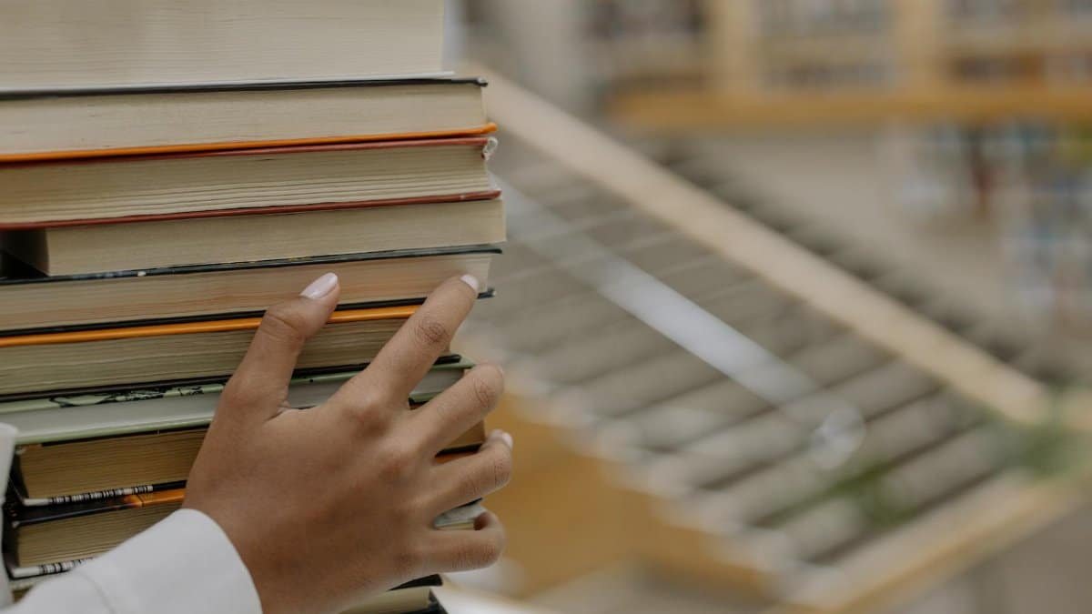 A hand holding a stack of books with bookshelves blurred in the background, depicting knowledge and learning.
