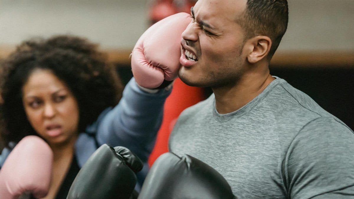 Side view of young muscular ethnic male trainer in sportswear and boxing gloves receiving heavy punch on face from serious young African American female during workout