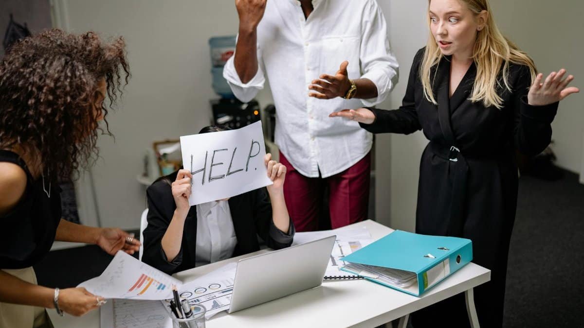 A group of employees in heated discussion, one holding a 'HELP' sign, indicates workplace stress.