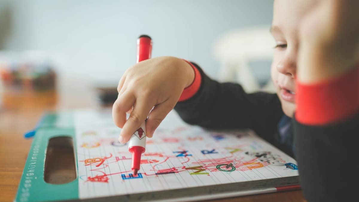 A young child using a marker to write letters in an educational workbook.
