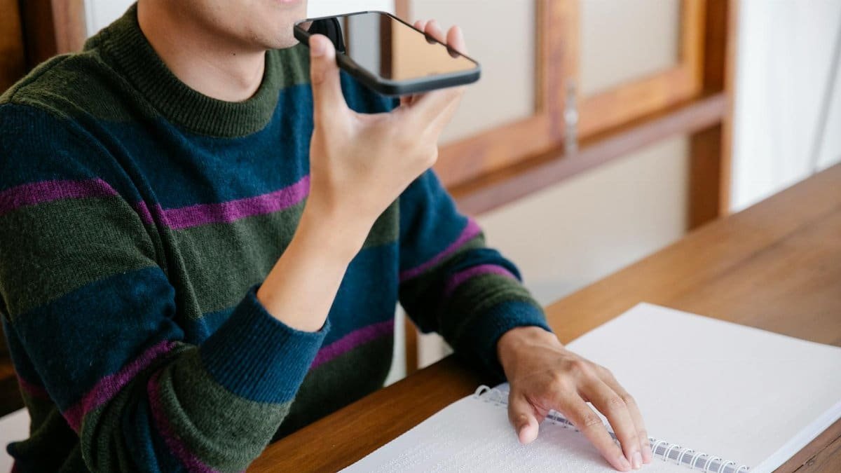 Adult man using smartphone for voice commands, smiling at home desk.