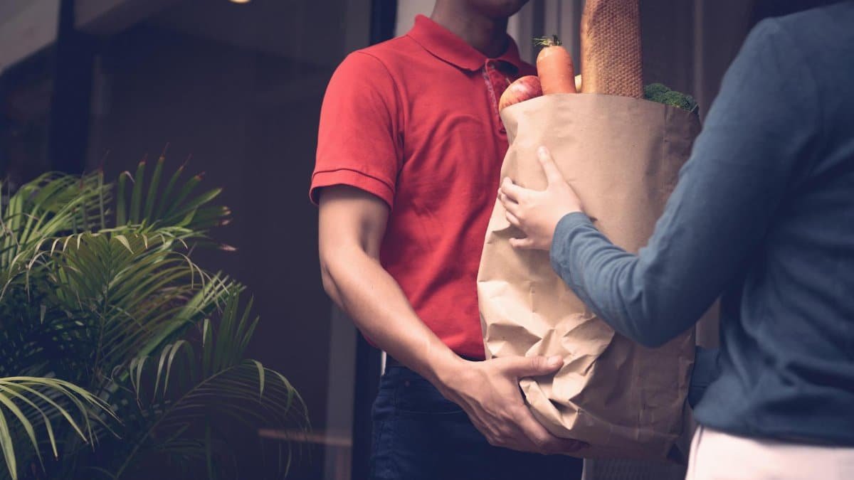A delivery person in red shirt delivers a grocery bag filled with food items to a customer at the door.
