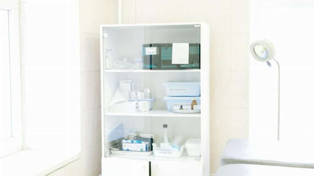 Modern white cabinet with cupboard with various medical instruments and shelves with medical materials near empty white bed near window and lamp in hospital
