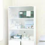 Modern white cabinet with cupboard with various medical instruments and shelves with medical materials near empty white bed near window and lamp in hospital