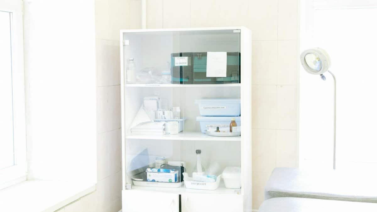 Modern white cabinet with cupboard with various medical instruments and shelves with medical materials near empty white bed near window and lamp in hospital