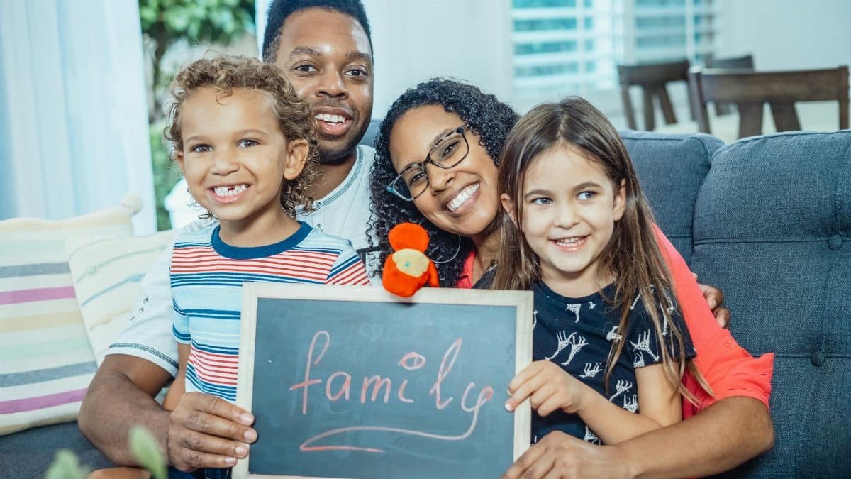 Smiling family of four indoors holding a blackboard with 'family' written on it.