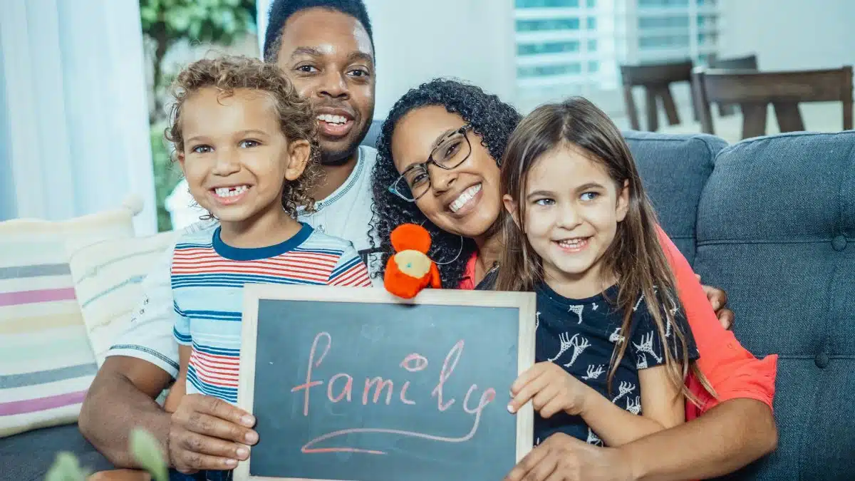 Smiling family of four indoors holding a blackboard with 'family' written on it.