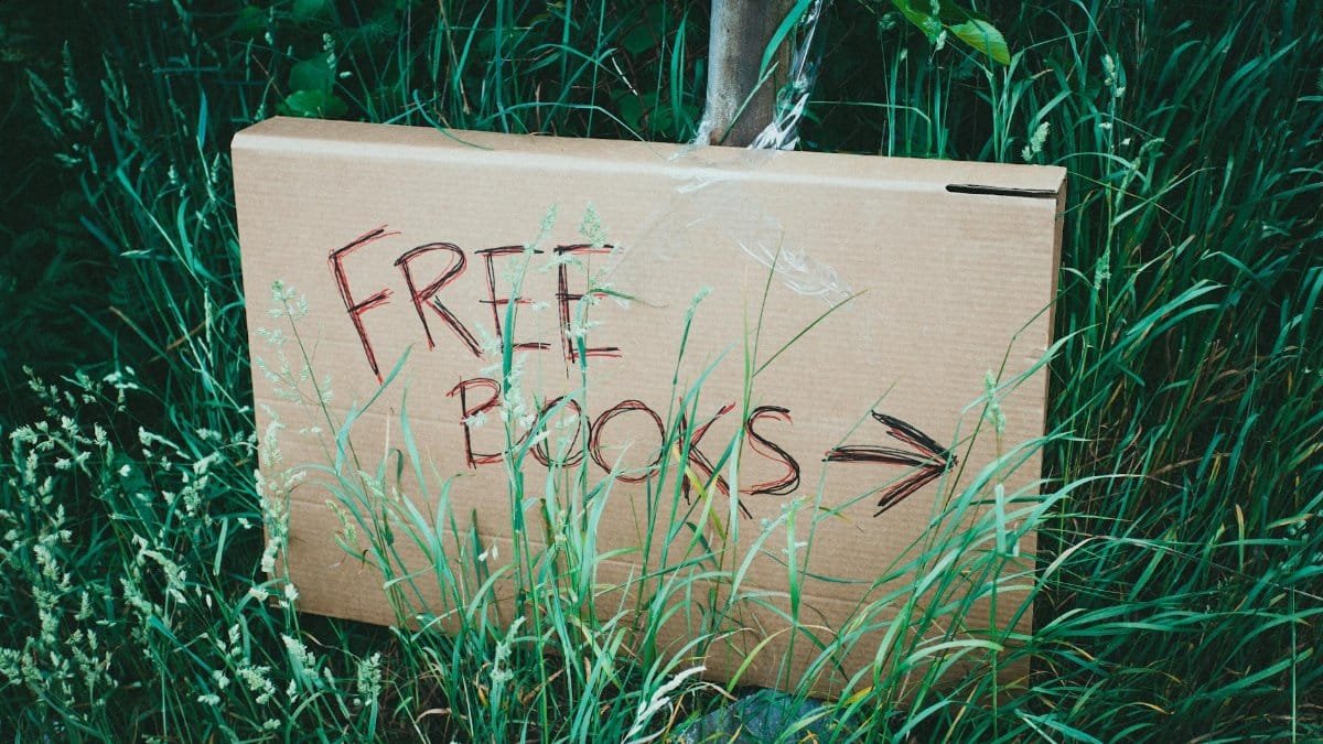 A cardboard sign with 'Free Books' handwritten, placed among green grass outdoors promoting book exchange.