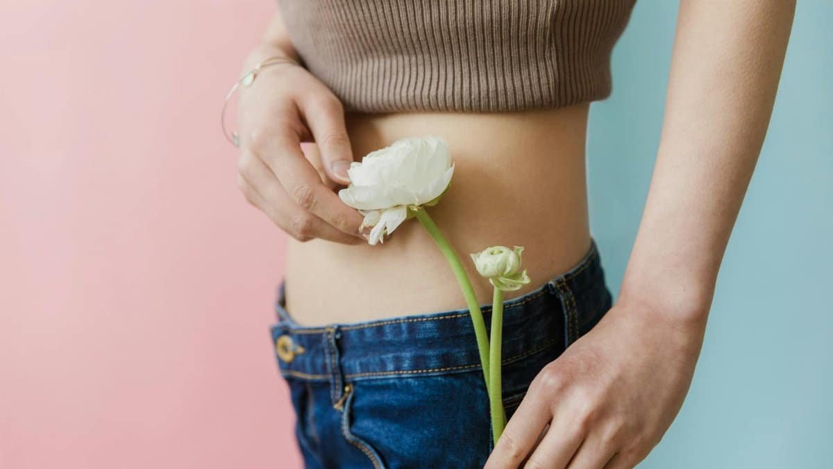 Fashionable woman in jeans and crop top holding a white flower against pink and blue backgrounds.
