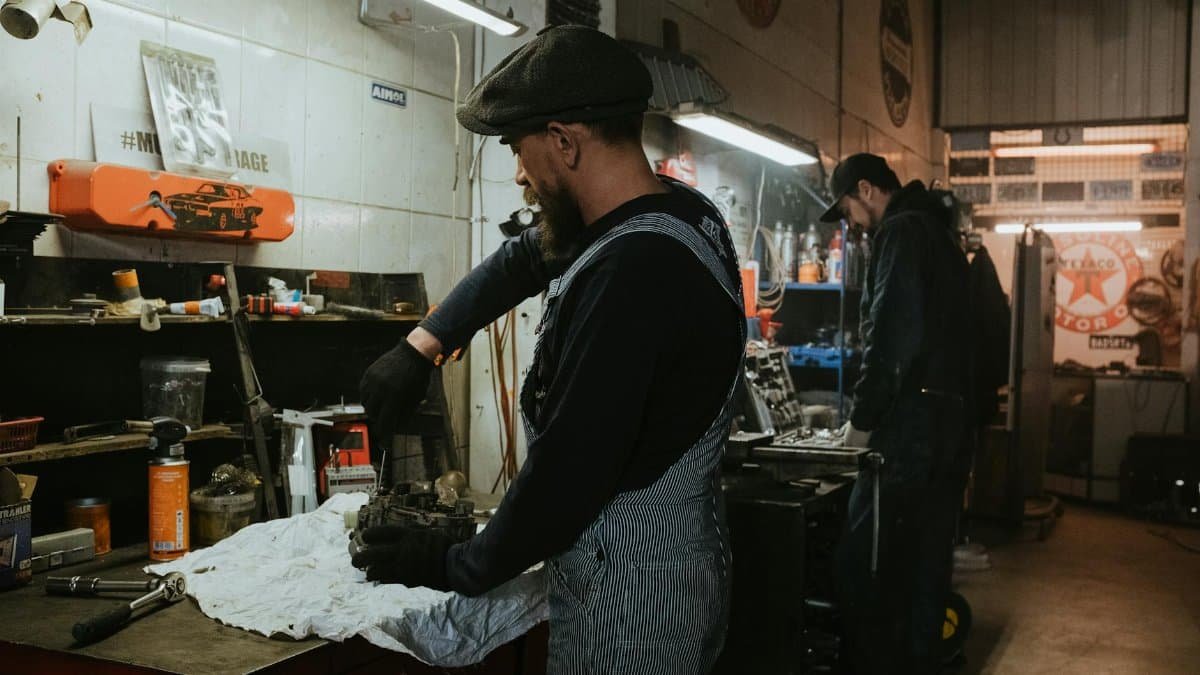 Two auto mechanics engaged in vehicle repair work inside a dimly lit garage.