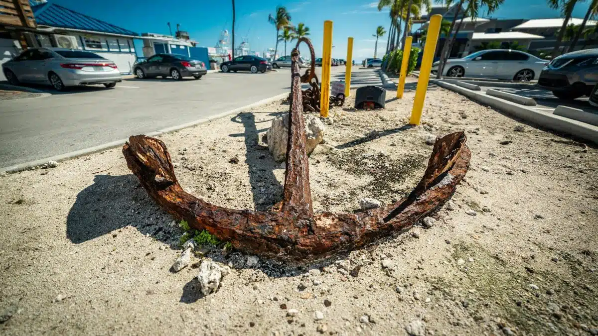 A weathered anchor on display in a sunny Florida parking lot with palm trees and cars.