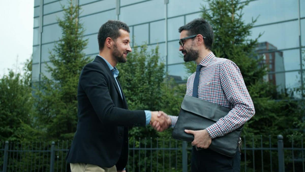 Two businessmen shaking hands outside a modern office building, symbolizing partnership.
