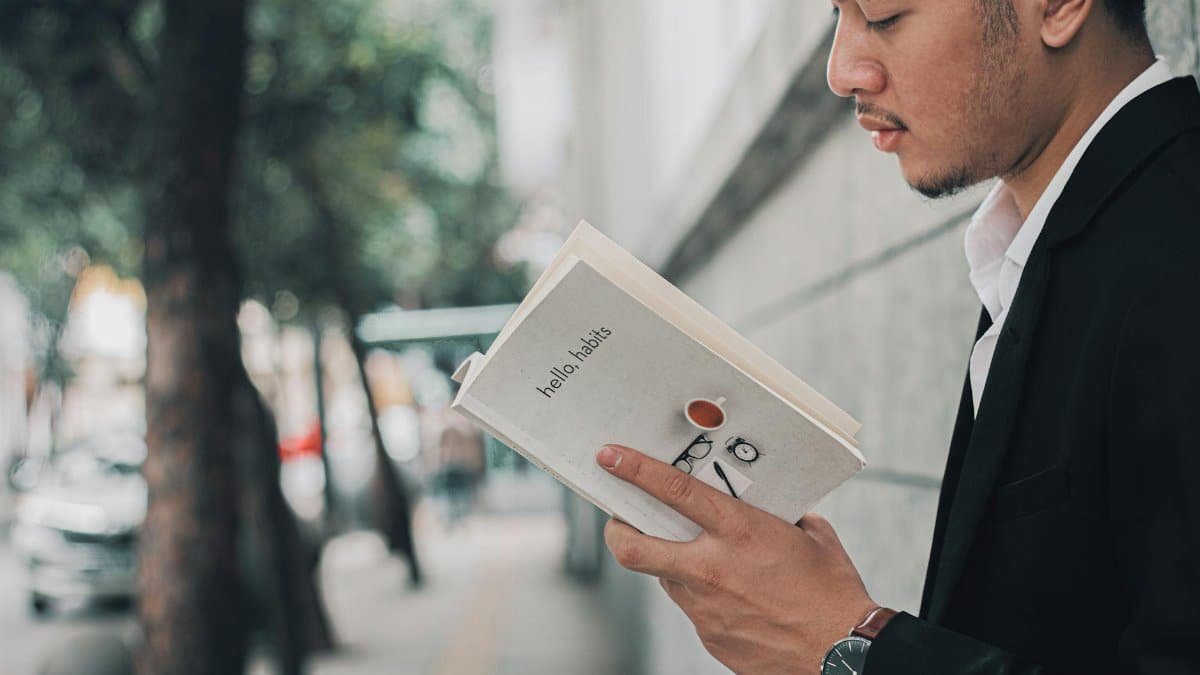 A man in a suit reading a book titled 'hello habits' on a city sidewalk.
