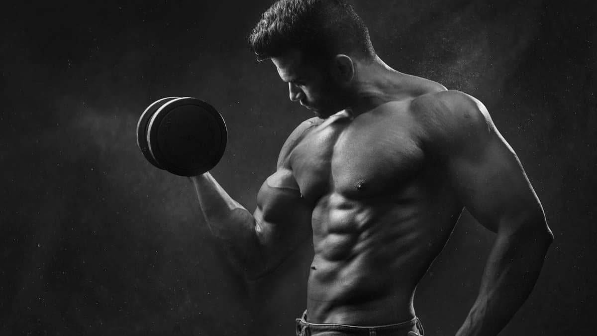 Powerful black and white portrait of a shirtless bodybuilder lifting dumbbells in the gym.