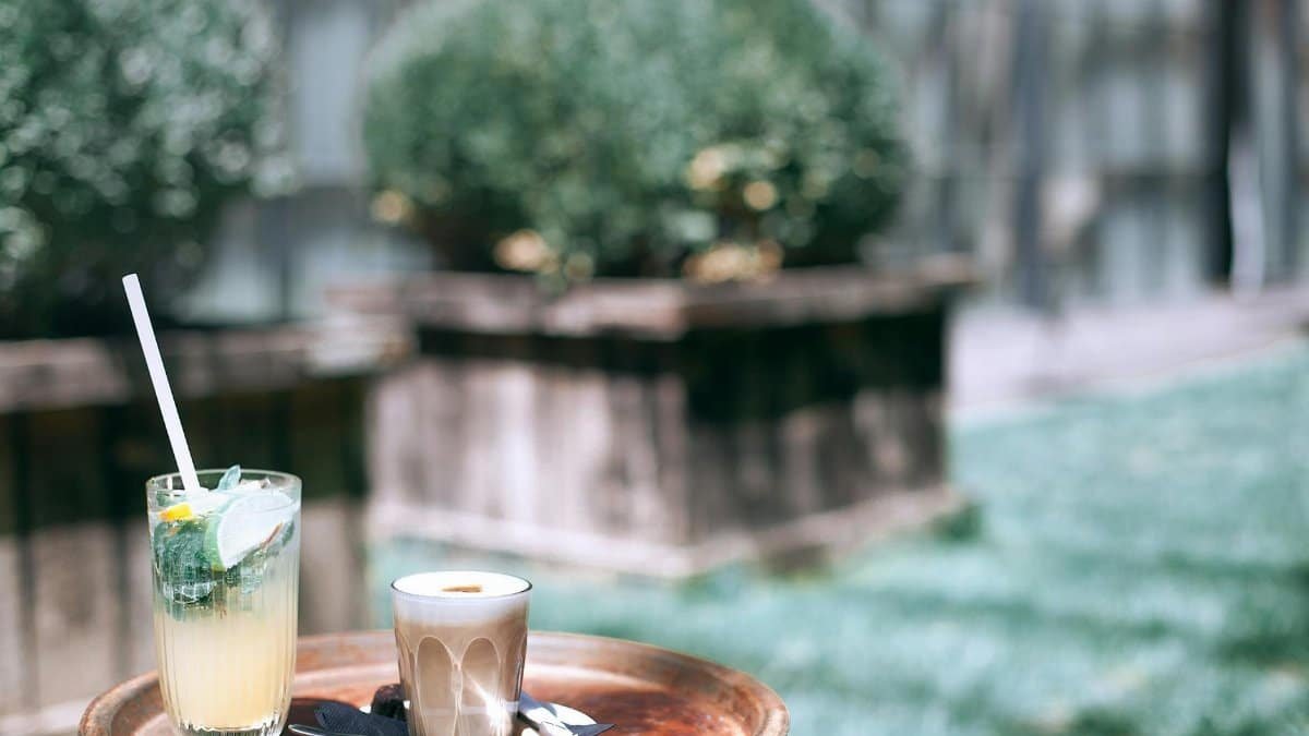 Round tray with glass of delicious cold cocktails and hot coffee placed on street against green bushes on blurred background