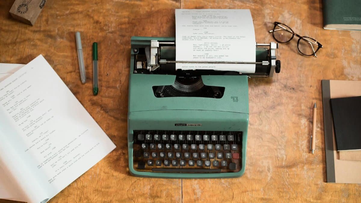 A vintage green typewriter with scripts surrounded by glasses and pens on a wooden table.