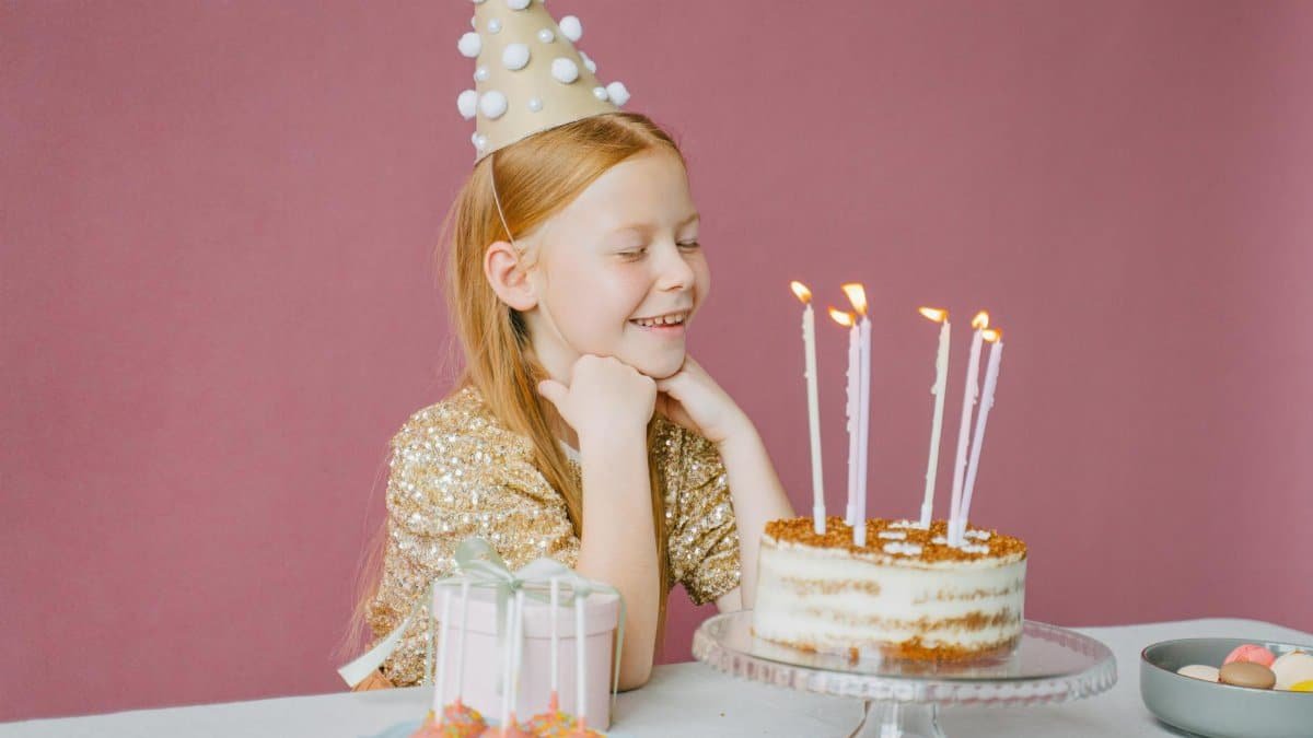 A young girl in a party hat smiles at her birthday cake with candles lit, ready to make a wish.
