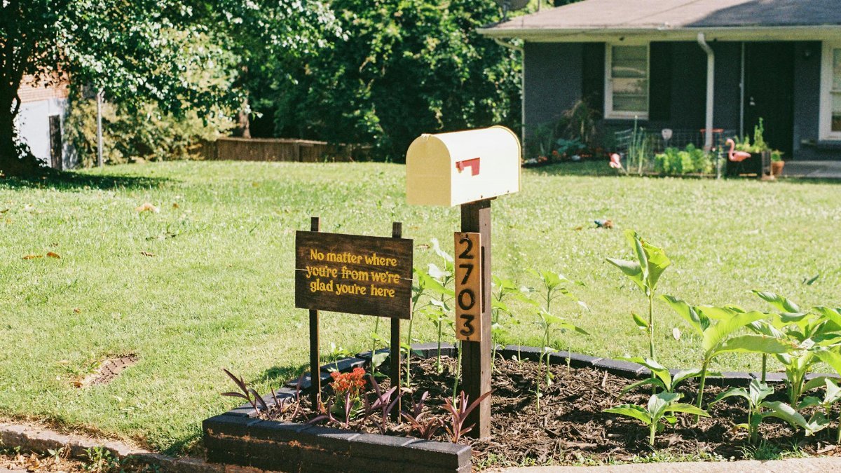 Yellow mailbox with a warm welcoming sign and plants in a suburban front yard.