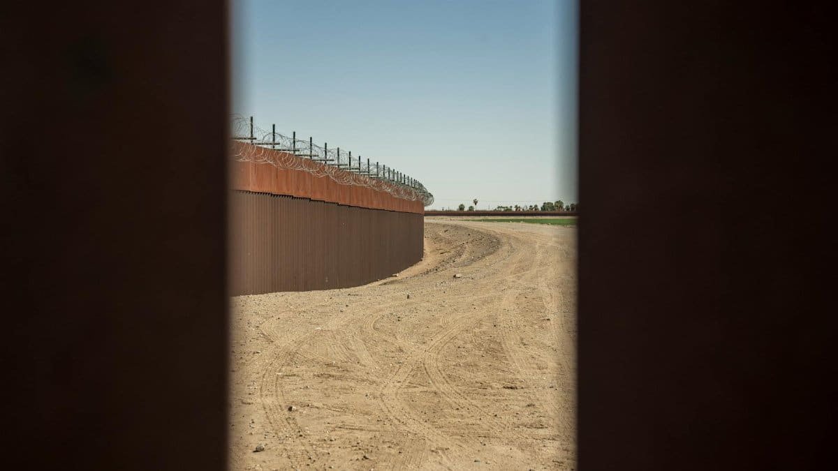 View of a barbed wire border wall structure alongside a dirt road under a clear blue sky.
