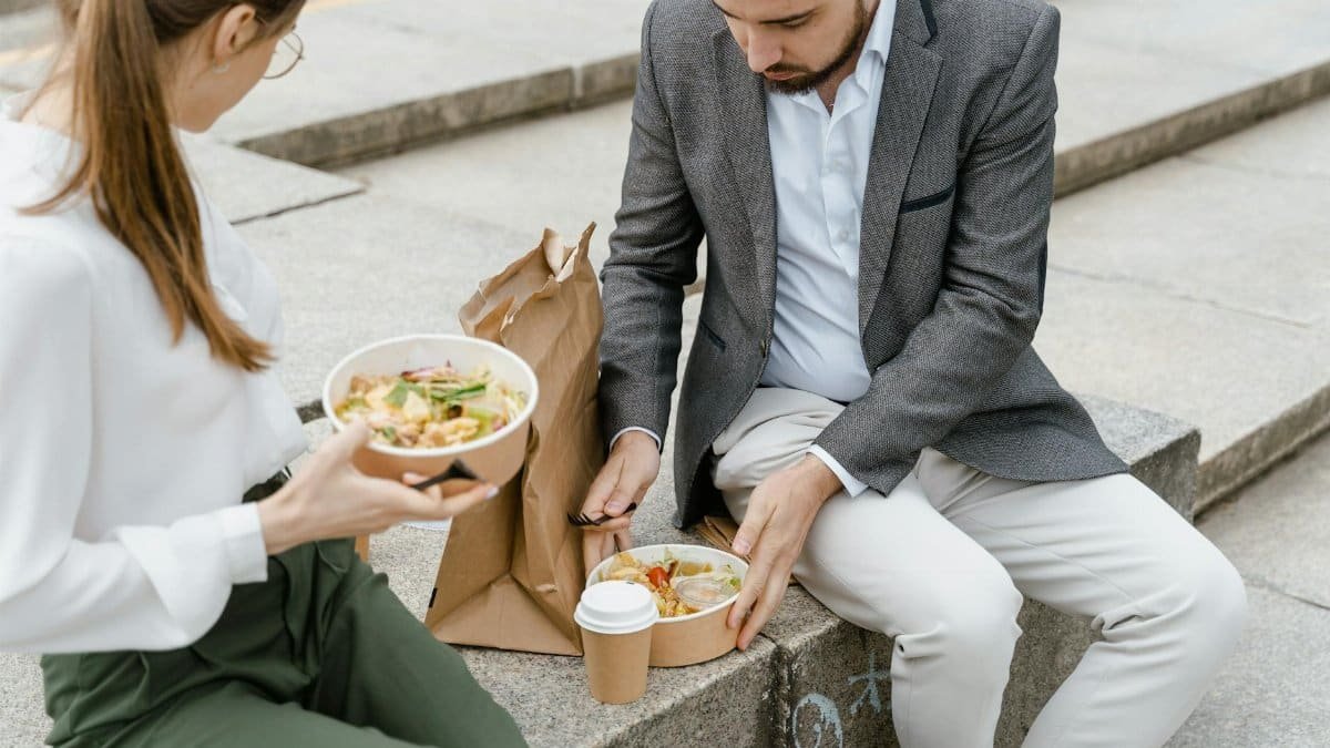 Man and woman enjoying takeaway lunch on stairs in a park. Casual outdoor meal with hot beverage and paper bags.
