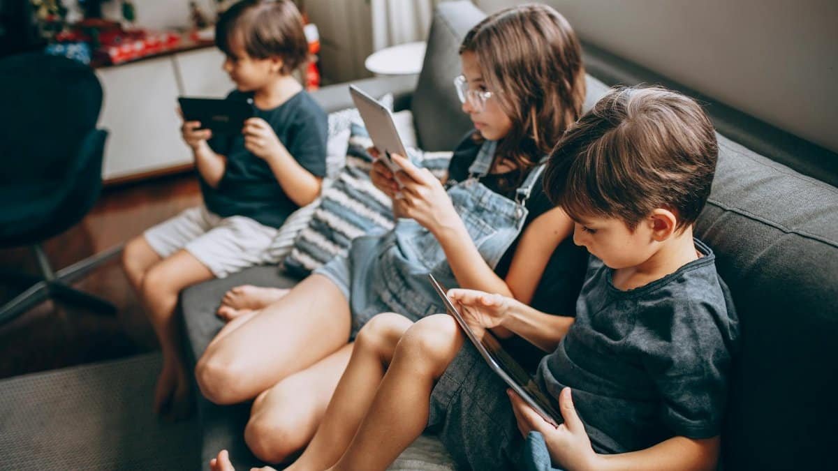 Three children using digital tablets while sitting together on a cozy sofa, enjoying technology indoors.