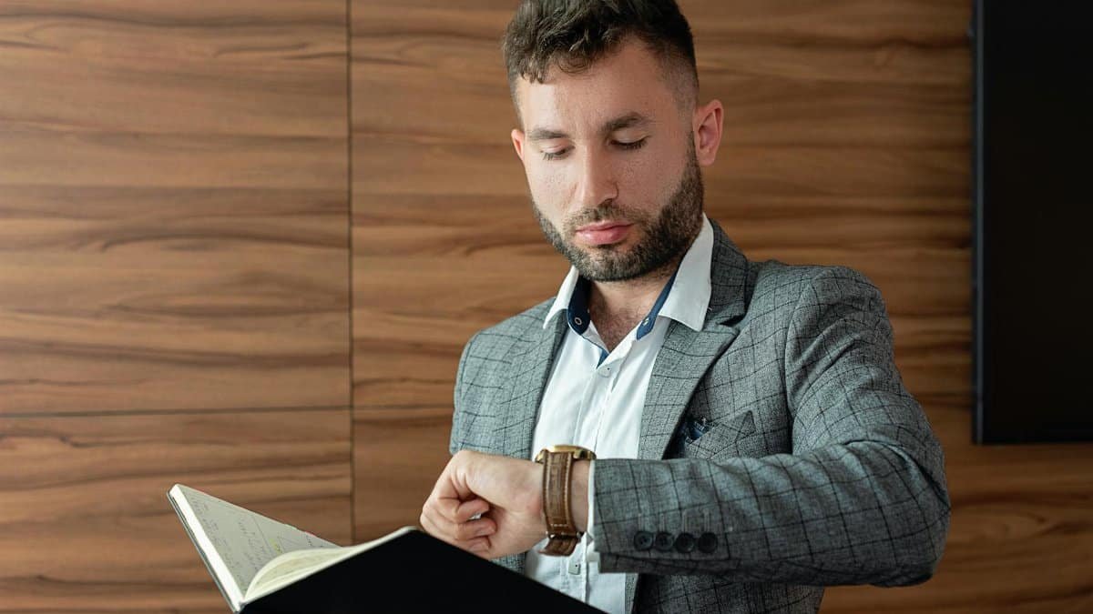 Young businessman in gray suit checking wristwatch while holding a notebook indoors.