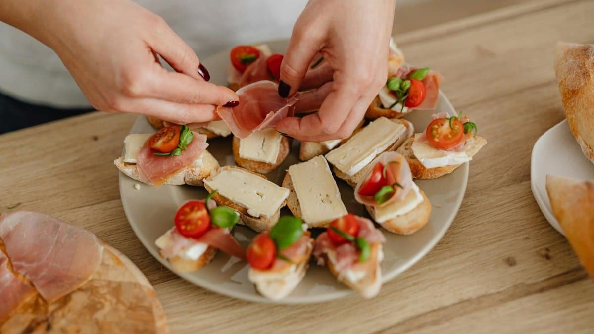 Close-up of hands preparing cheese and prosciutto appetizers on a wooden platter.