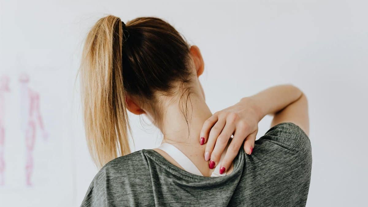 Back view of slim blond female in casual wear with manicure indicating pain spot on back while sitting in front of white wall with paper drawing in clinic